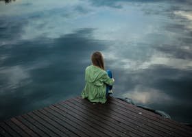 A woman sits on a wooden dock, reflecting by a calm lake under a cloudy sky.