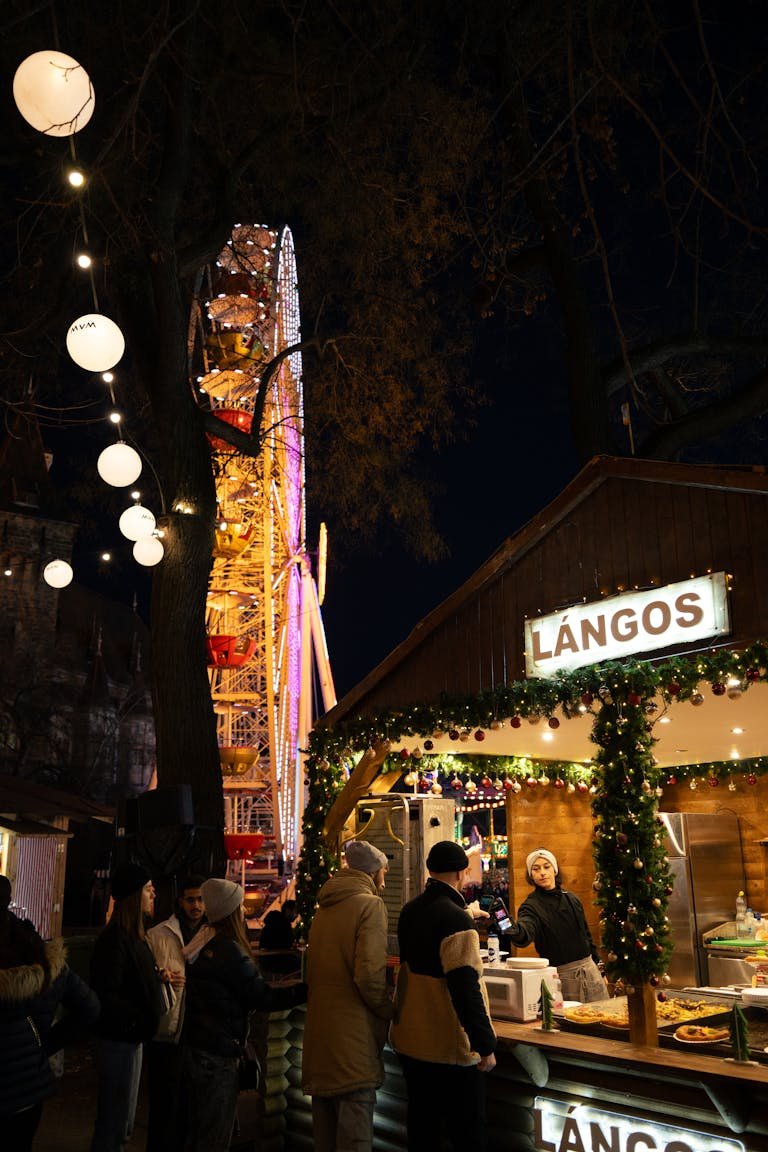 Night view of a Langos stall at Budapest Christmas market with Ferris wheel lights. Living Costs Budapest